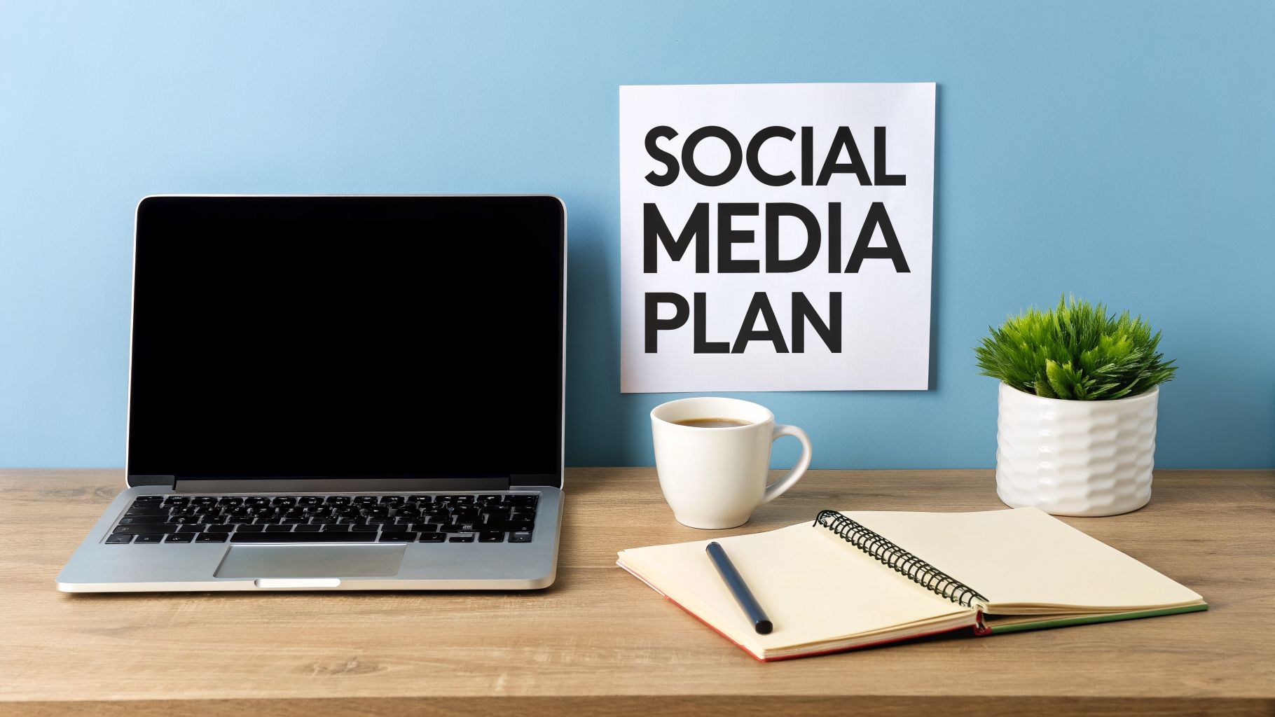 Ruang kerja dengan laptop, kopi, buku catatan, tanaman, dan tanda 'SOCIAL MEDIA PLAN'.